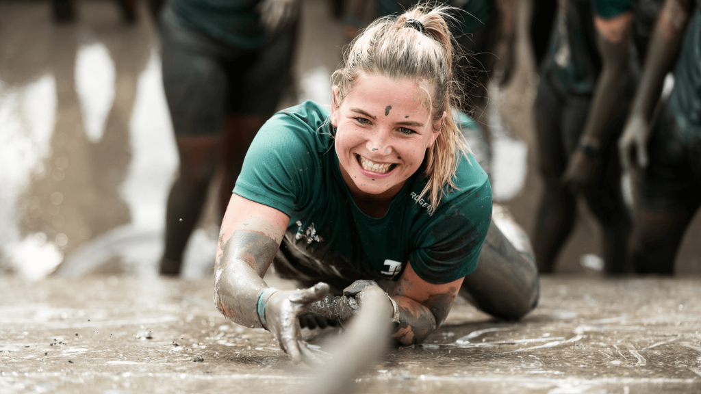 Een vrouw die lachend onder de modder zit en zichzelf met een touw over een muur hijst tijdens de Strong Viking Obstacle Run.