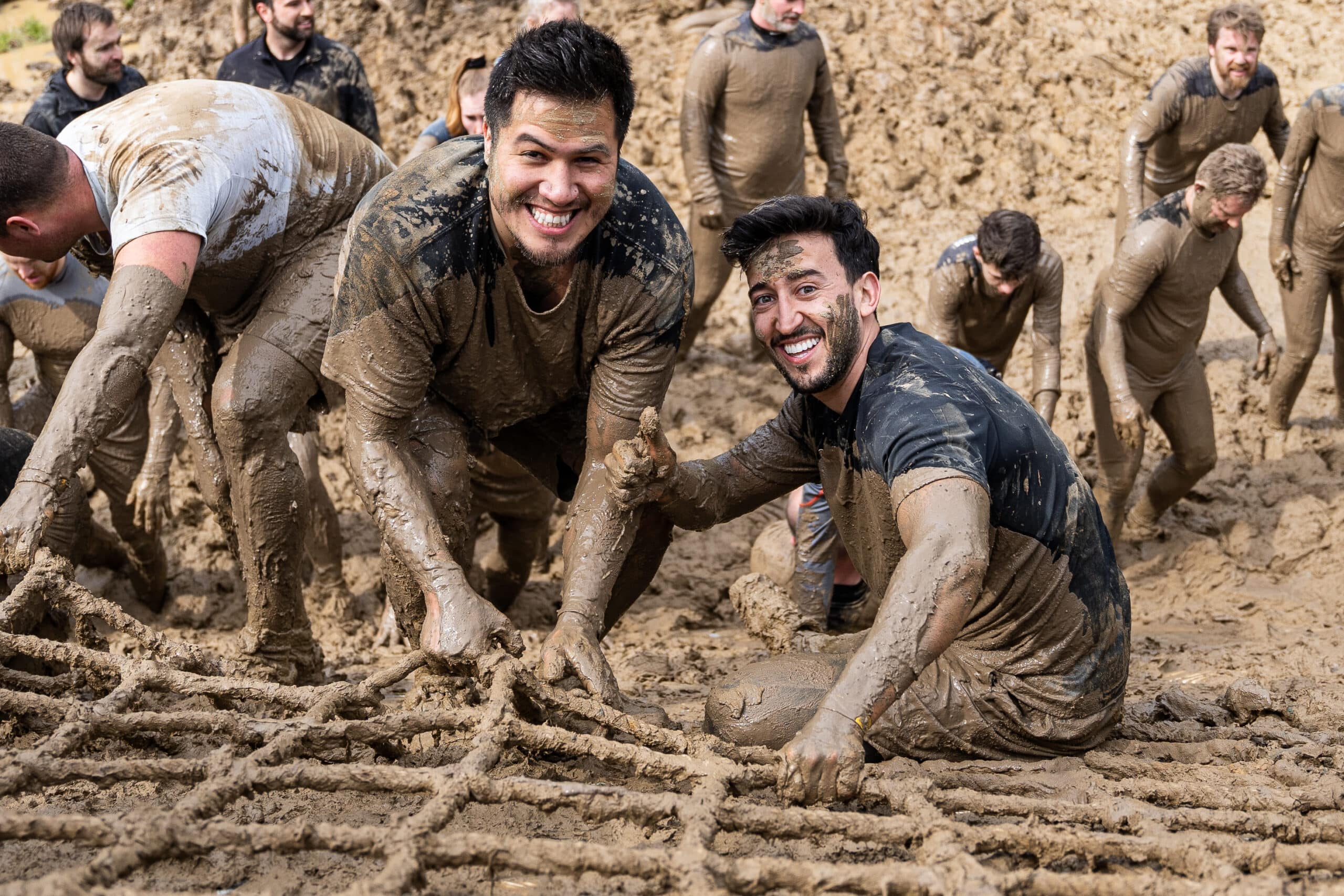 Two boys smiling at the camera and covered in mud while climbing up a net. Photo for getting mud out of your clothes after an obstacle run.